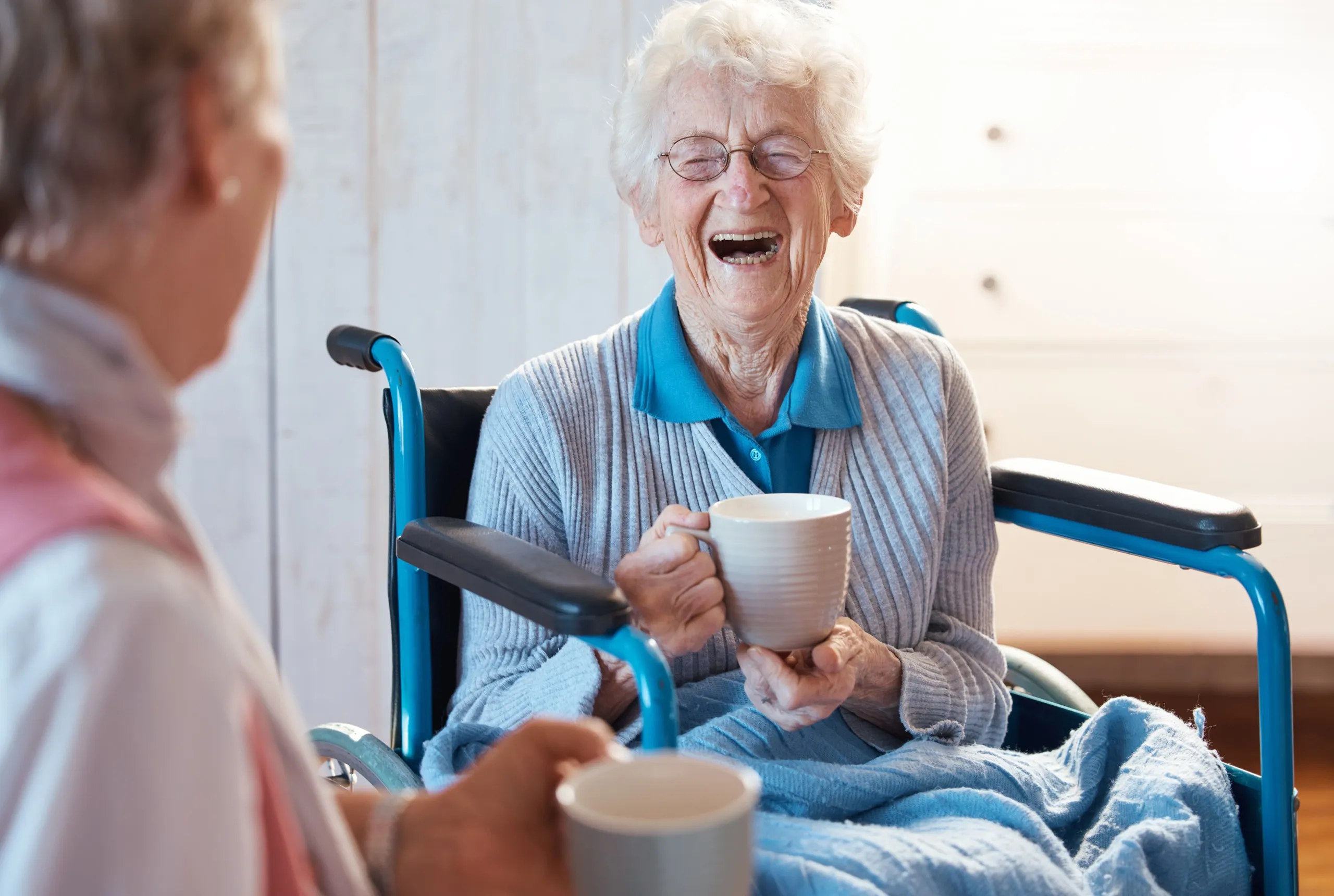 Lachende Seniorin im Rollstuhl beim Kaffee mit einer Pflegekraft