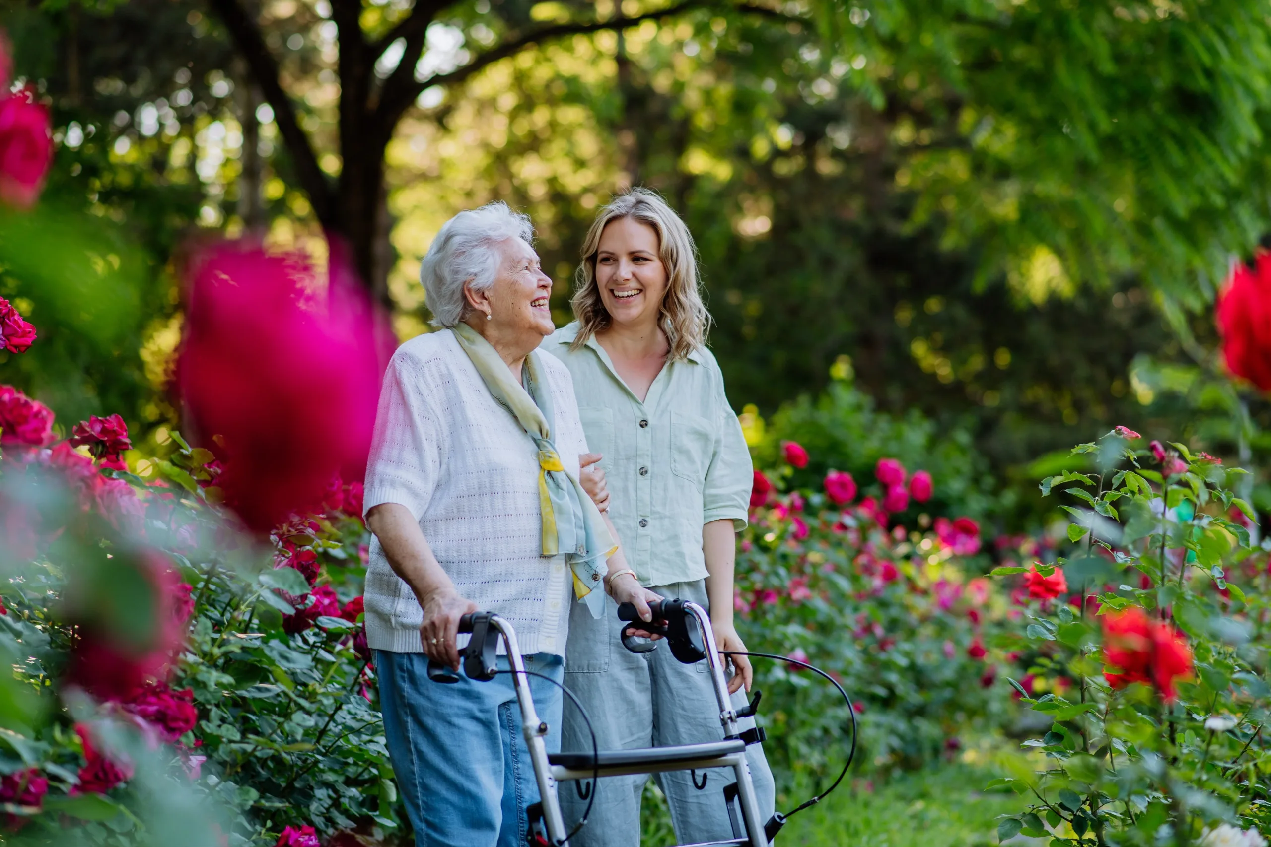 Pflegekraft begleitet eine Seniorin mit Rollator durch einen Rosengarten