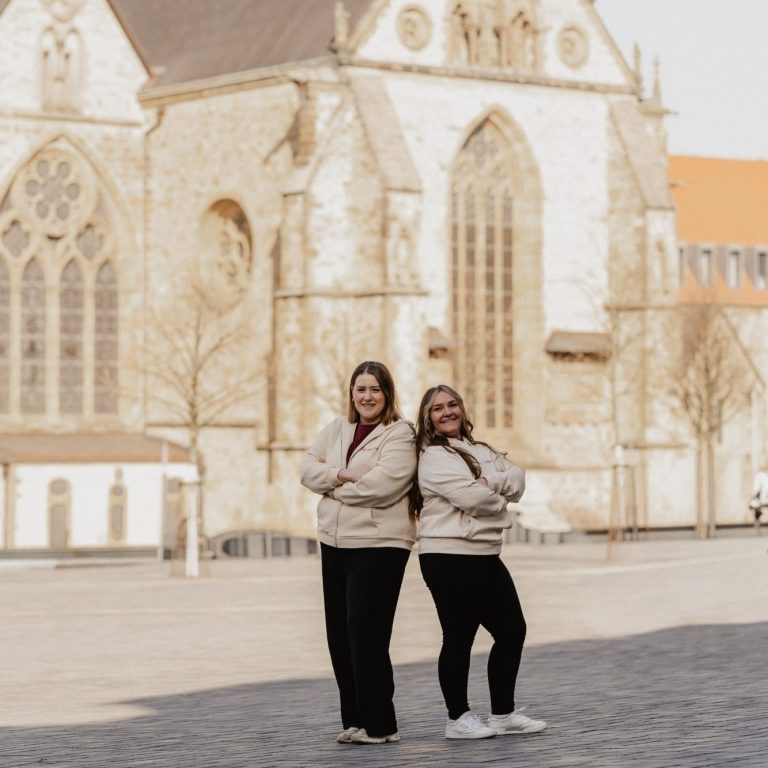 Zwei Frauen stehen vor einer alten Kirche auf einem Platz.