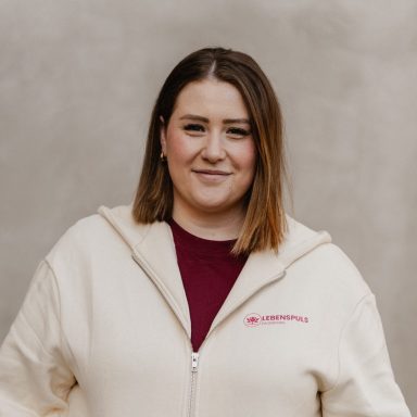 Junge Frau mit schulterlangem, braunem Haar in einer hellen Kapuzenjacke, lächelnd.