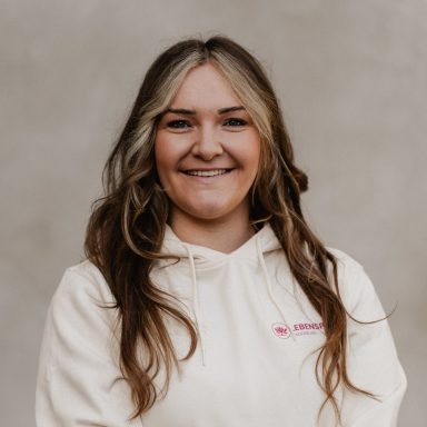 Lächelnde Frau mit langem, gewelltem Haar in einem weißen Hoodie vor neutralem Hintergrund.