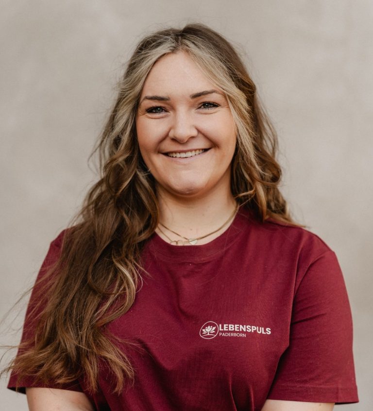Junge Frau mit langen, lockigen Haaren, die ein rotes T-Shirt trägt und lächelt freundlich.