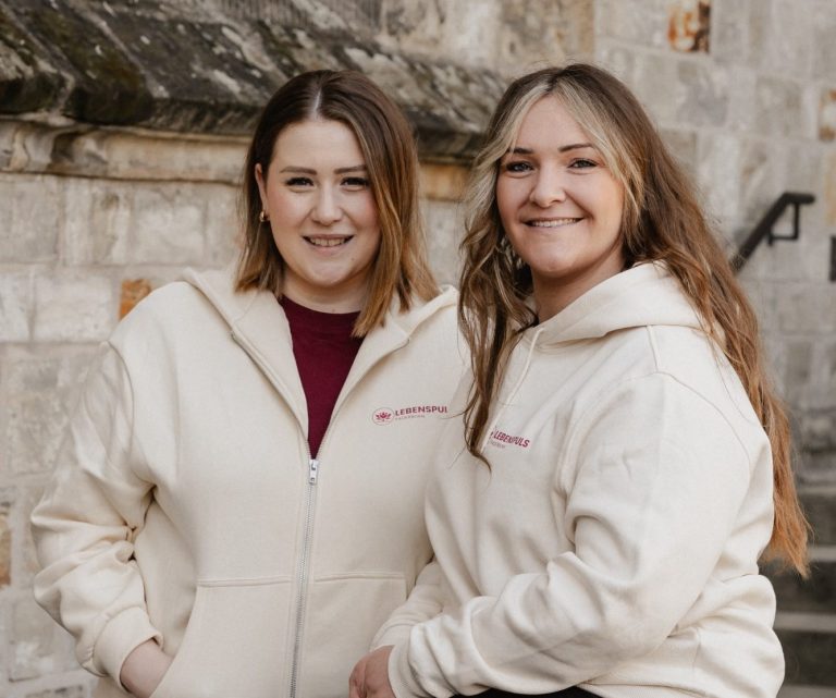 Zwei Frauen in beigen Kapuzenpullovern lächeln vor einer steinernen Wand.